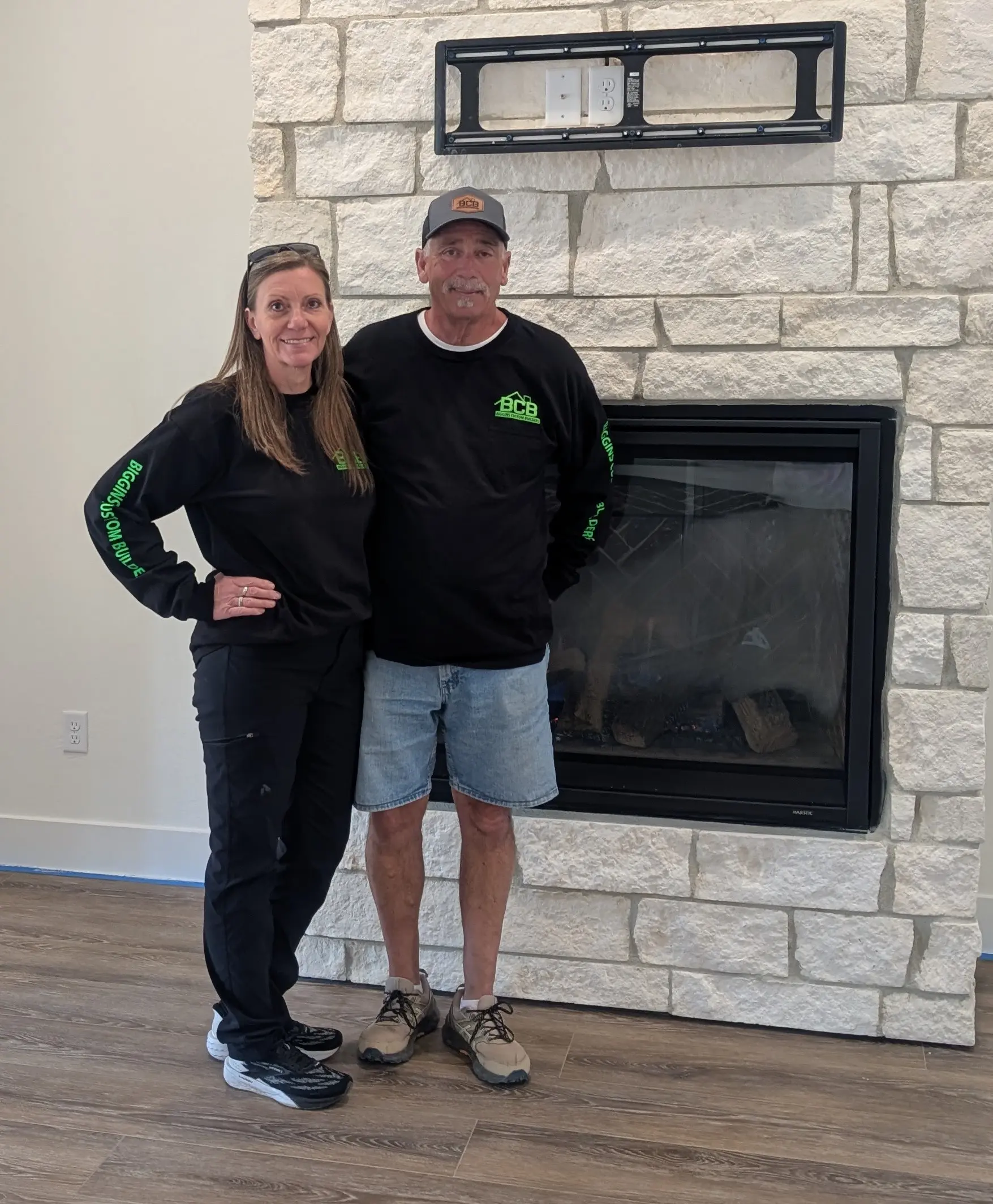 A smiling couple stands in front of a stone fireplace inside a cozy home.