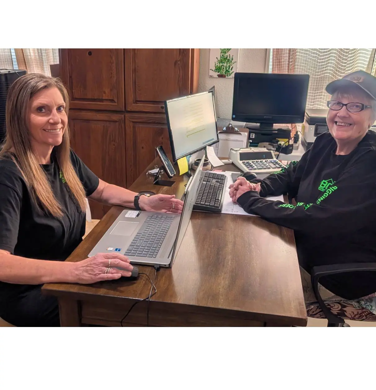 Two people smiling while working on laptops at a wooden table.