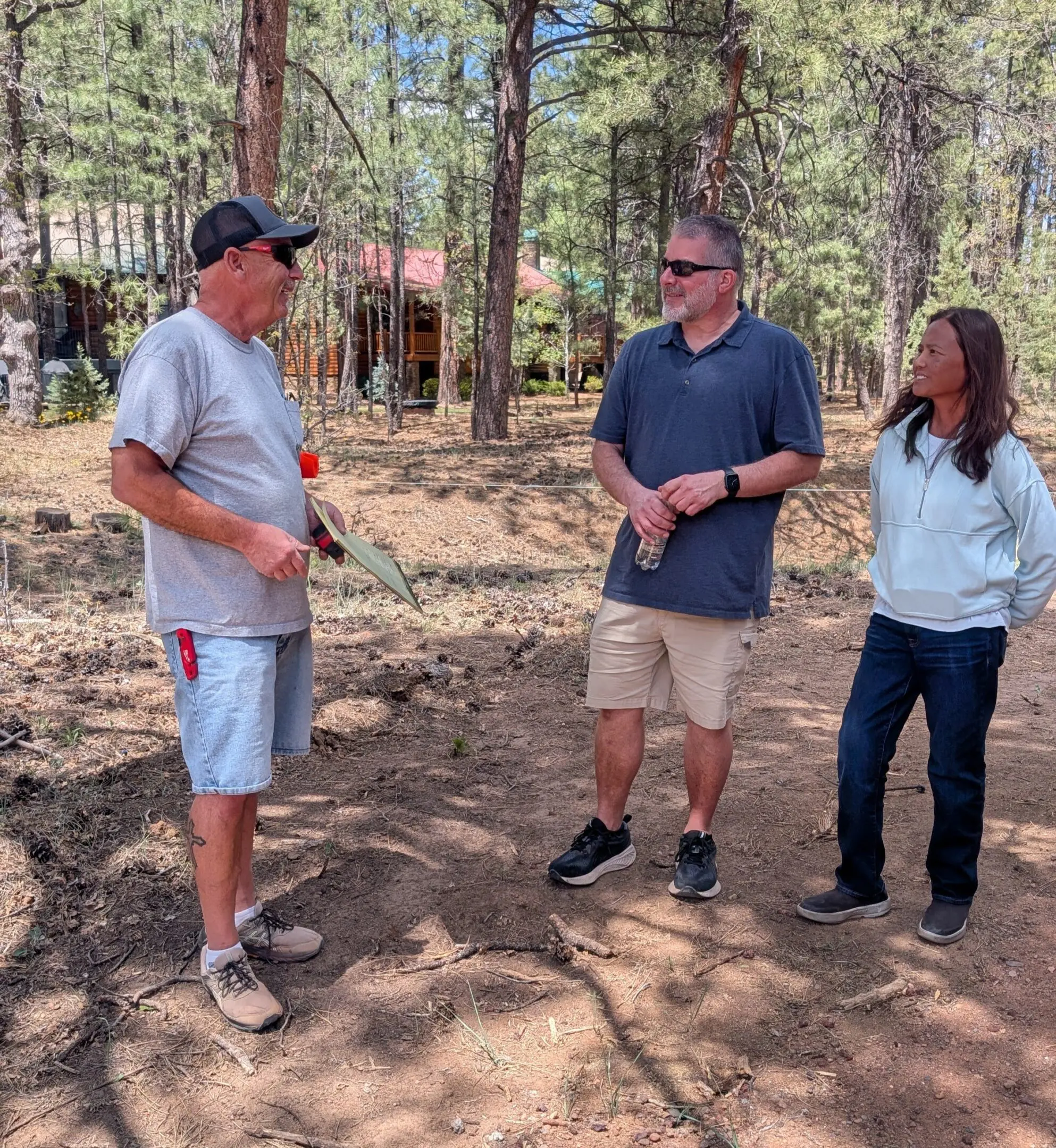 Three people standing and talking in a wooded outdoor area.